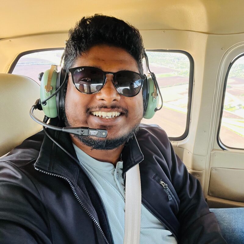 Nidhin smiling in the cockpit of a small plane, wearing a headset and sunglasses